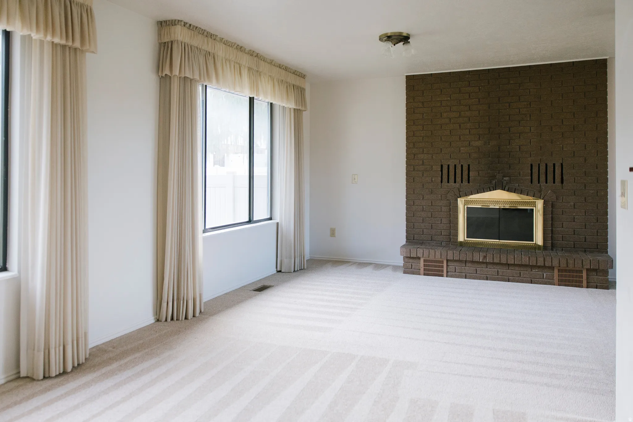 Spacious interior room featuring a prominent brick-clad fireplace with a brass firebox, flanked by large windows with drapery