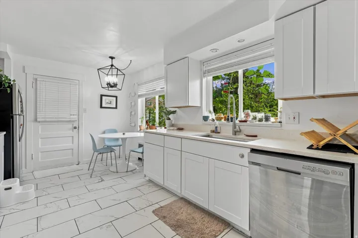 White shaker cabinetry, light-toned countertops, tile flooring, stainless steel dishwasher, and a window over the sink