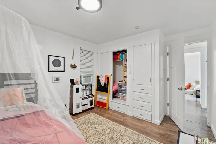 Bright bedroom featuring wood-finish flooring, white built-in cabinetry with drawer storage, a walk-in closet with mirrored doors and interior shelving, and a ceiling-mounted light fixture