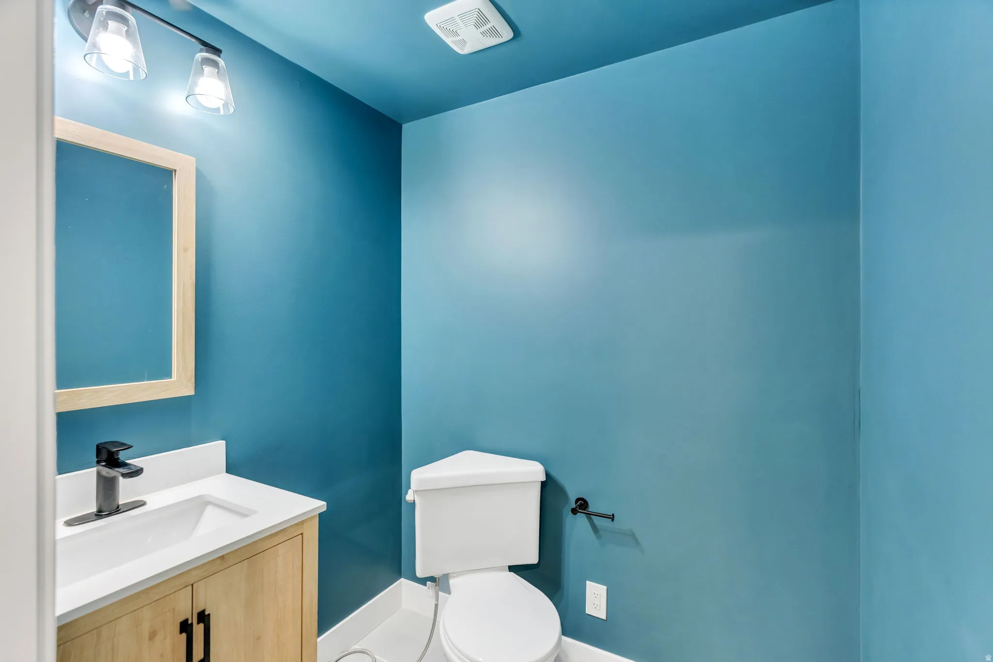 Powder room featuring a wood-finish vanity with a white countertop and integrated sink, matte black faucet, and rectangular framed mirror