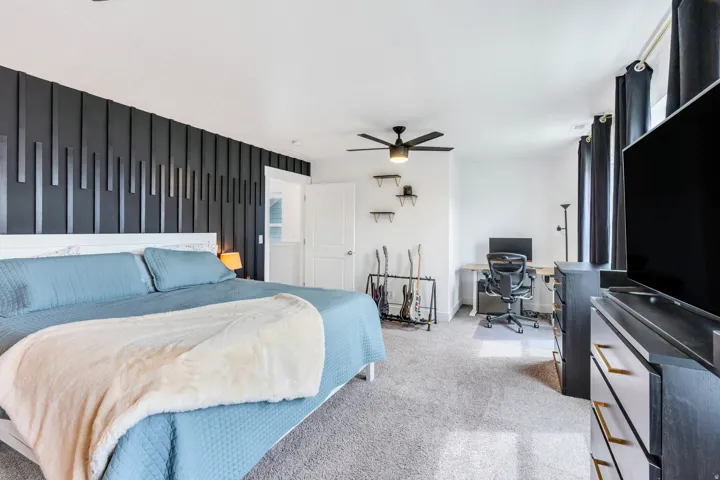 Spacious bedroom featuring a distinctive dark accent wall, light-toned carpet flooring, a ceiling fan with integrated lighting, and multiple windows with dark draperies