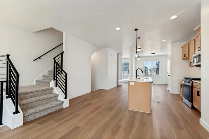 Open-concept living space featuring wood-finish flooring, a modern kitchen island with pendant lighting, light wood cabinetry with dark hardware, a stainless steel range, and a staircase with contemporary black metal railings