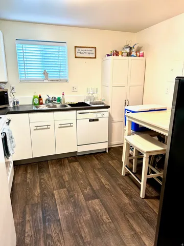 Kitchen featuring wood-finish flooring, white cabinetry with silver-tone hardware, a window with blinds, a built-in dishwasher, and a tall pantry cabinet