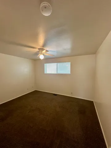 Carpeted room with a ceiling fan, a single window with horizontal blinds, and a recessed floor vent