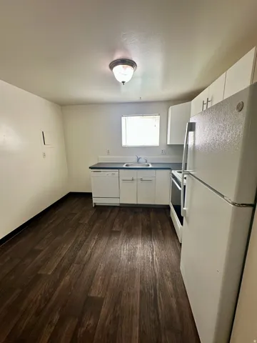 Modern kitchen space featuring wood-finish flooring, white cabinetry, dark countertops, a stainless steel sink, and white appliances