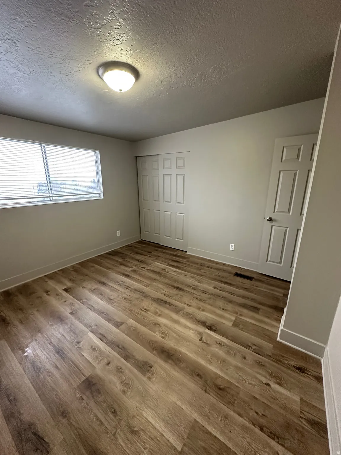 Spacious room featuring wood-finish flooring, a contemporary ceiling light fixture, a window with blinds, and white bi-fold closet doors