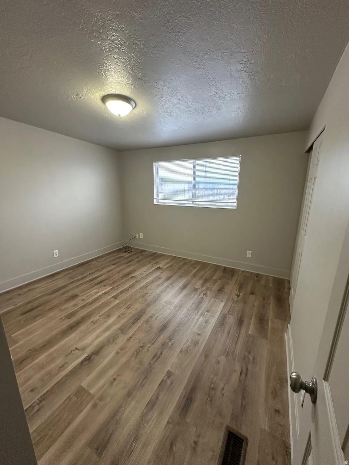 Spacious room featuring wood-finish flooring, neutral-toned walls, a single window with blinds, and a flush-mount ceiling light fixture