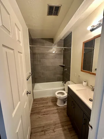 Bathroom featuring wood-finish flooring, a white vanity with a rectangular sink, a toilet, a shower-over-tub combination with dark gray subway tile surround, and an overhead light fixture