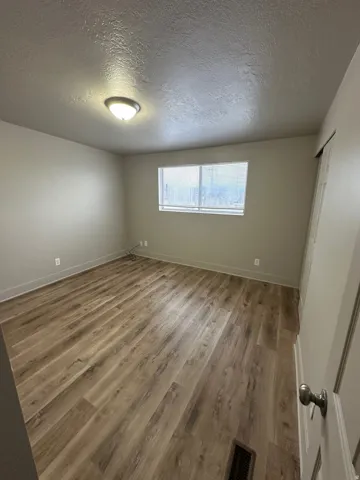 Spacious room featuring wood-finish flooring, neutral-toned walls, a single window with blinds, and a flush-mount ceiling light fixture