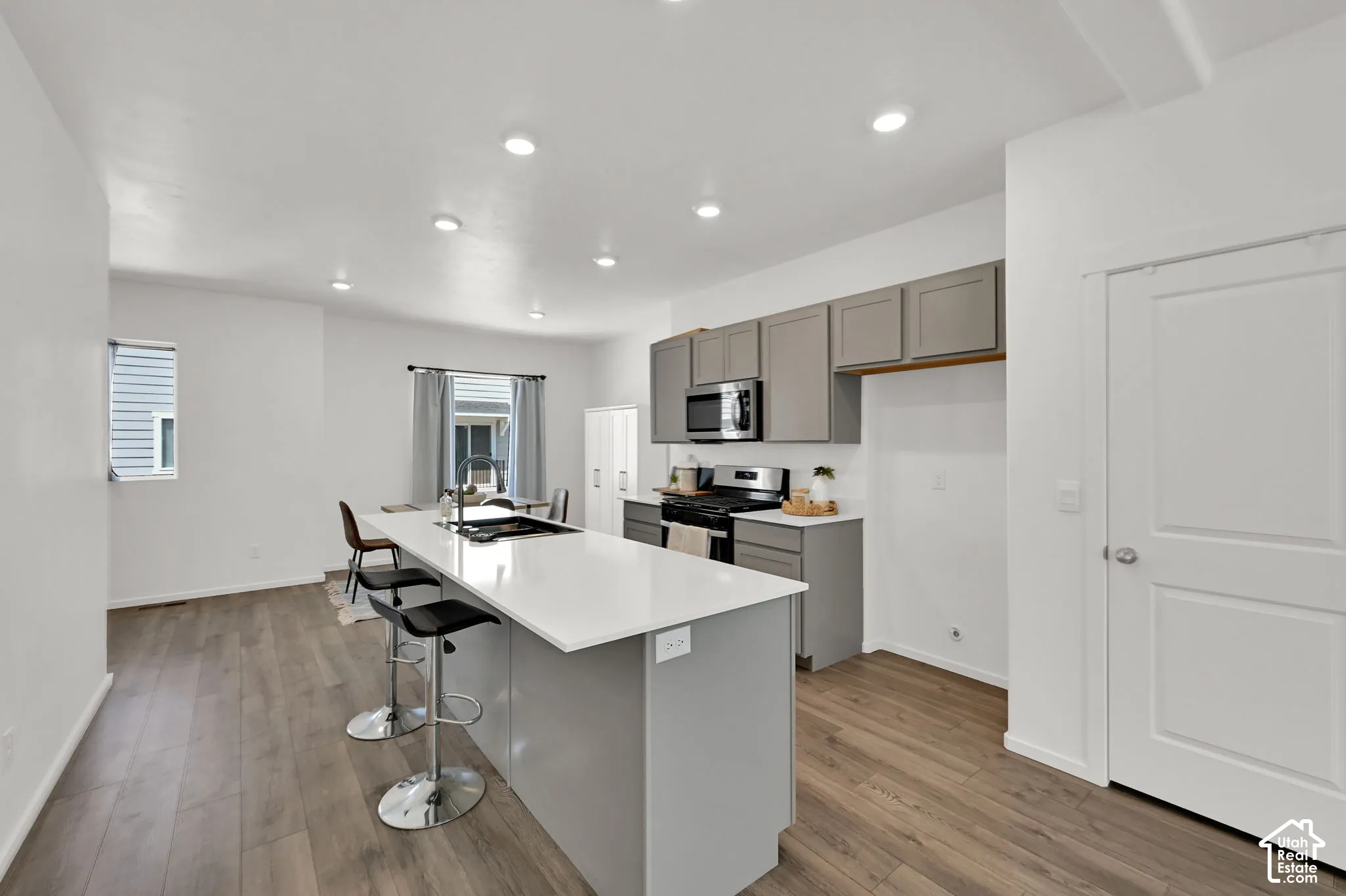 Open-concept kitchen featuring an island with a built-in sink, stainless steel appliances, grey cabinetry, wood-finish flooring, and recessed lighting