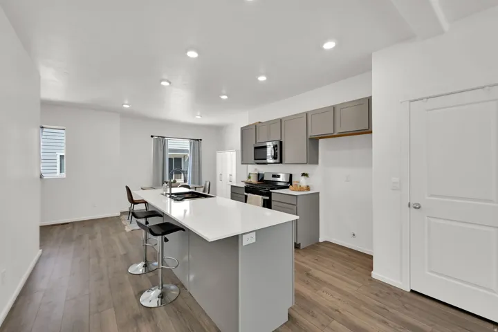 Open-concept kitchen featuring an island with a built-in sink, stainless steel appliances, grey cabinetry, wood-finish flooring, and recessed lighting