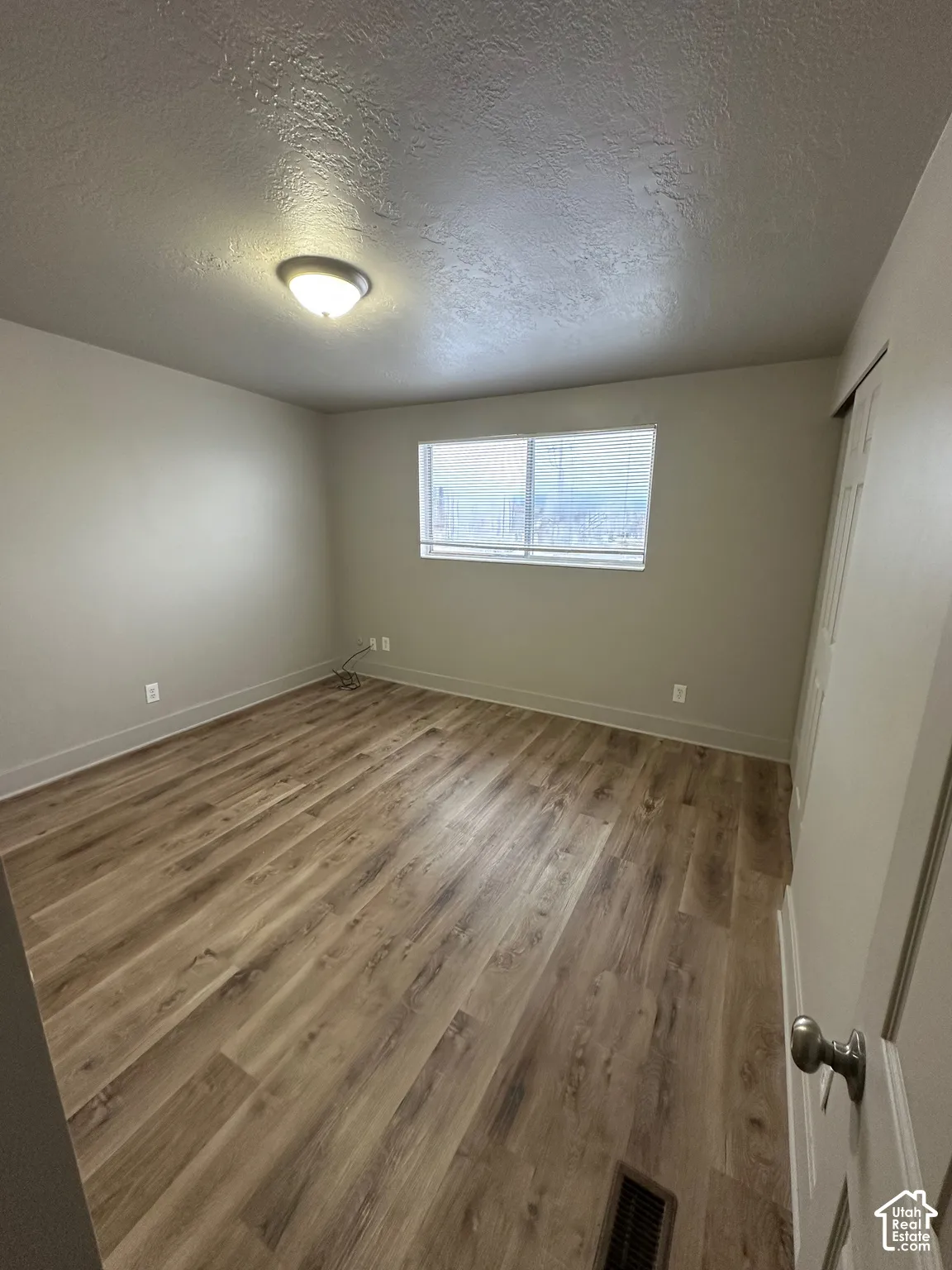 Spacious room featuring wood-finish flooring, neutral-toned walls, a single window with blinds, and a flush-mount ceiling light fixture