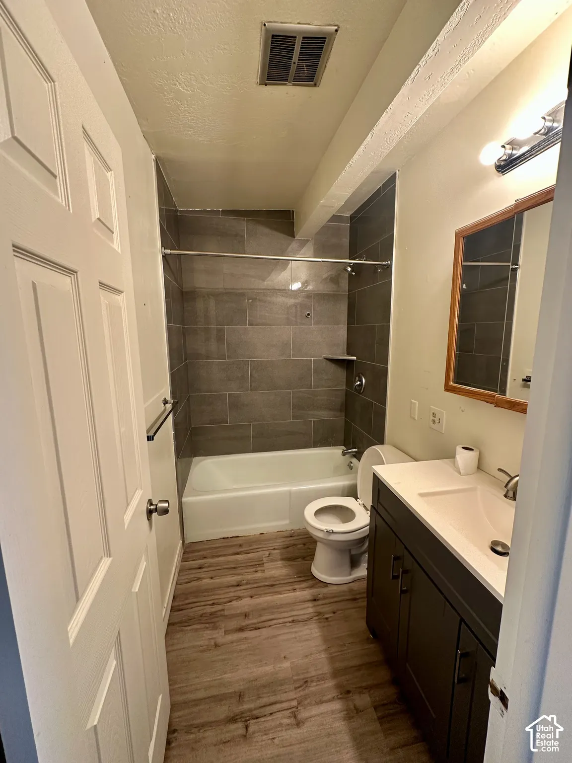 Bathroom featuring wood-finish flooring, a white bathtub with dark gray subway tile surround, a built-in shower niche, a white toilet, and a dark wood vanity with a white countertop and integrated sink