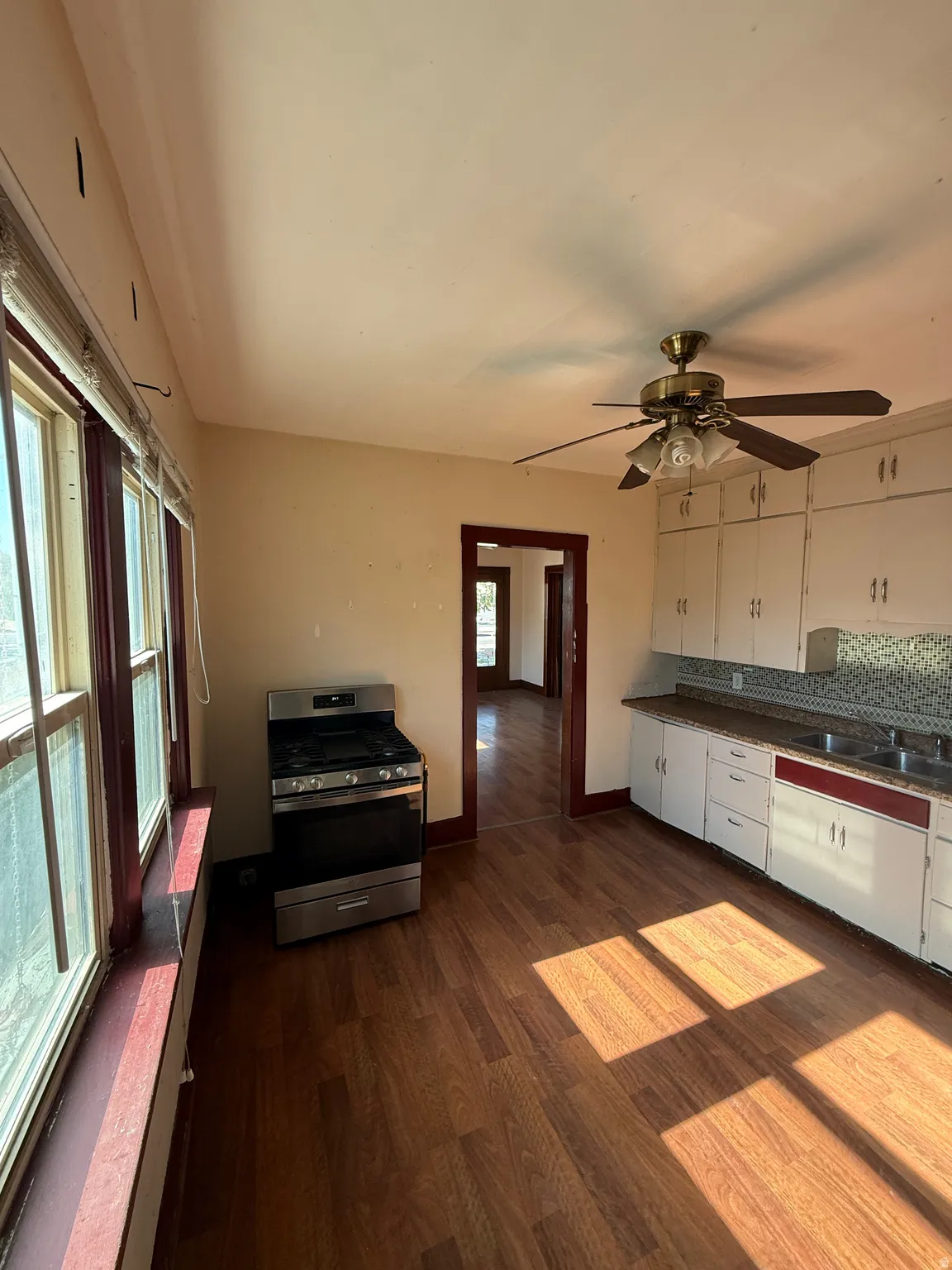 Kitchen featuring wood-finish flooring, white cabinetry with red trim, a stainless steel gas range, a brass-finish ceiling fan, and multiple windows