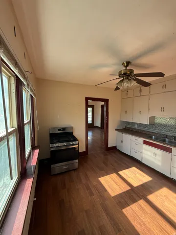 Kitchen featuring wood-finish flooring, white cabinetry with red trim, a stainless steel gas range, a brass-finish ceiling fan, and multiple windows