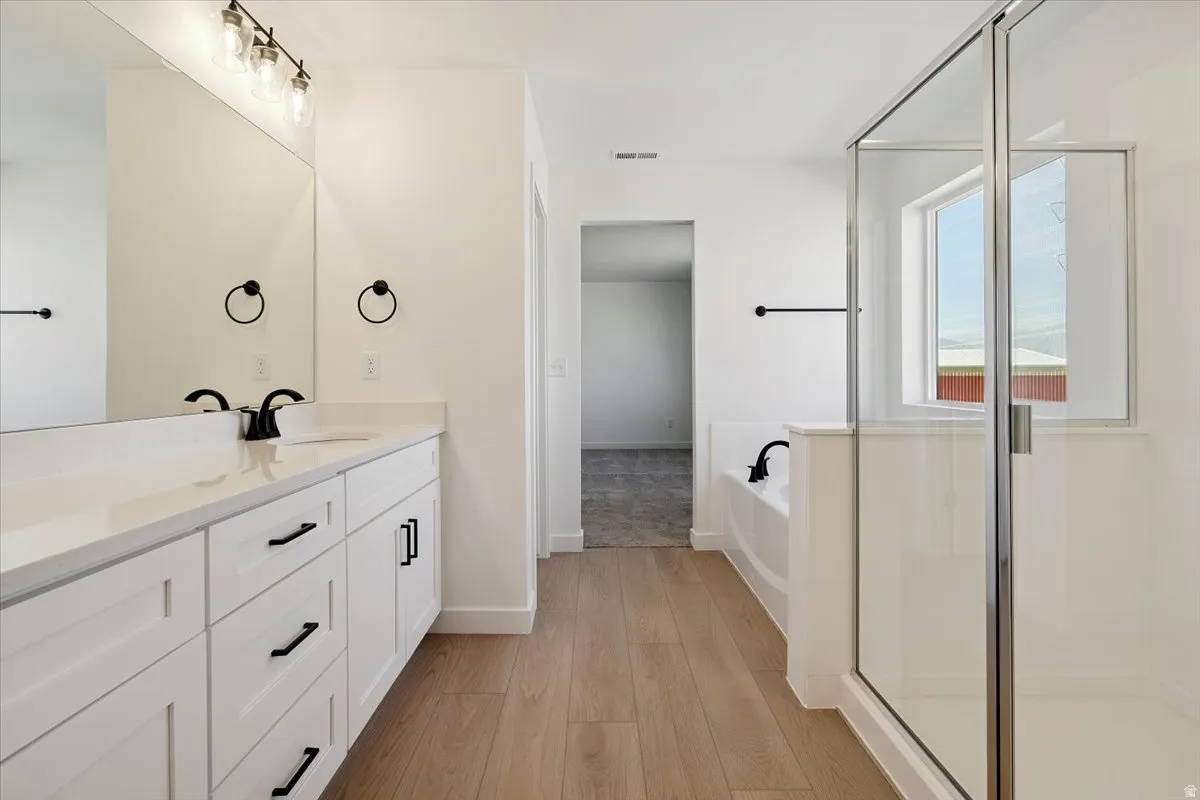 Spacious bathroom featuring a dual vanity with white cabinetry, a large mirror, a separate glass-enclosed shower, a built-in tub, and wood-finish flooring