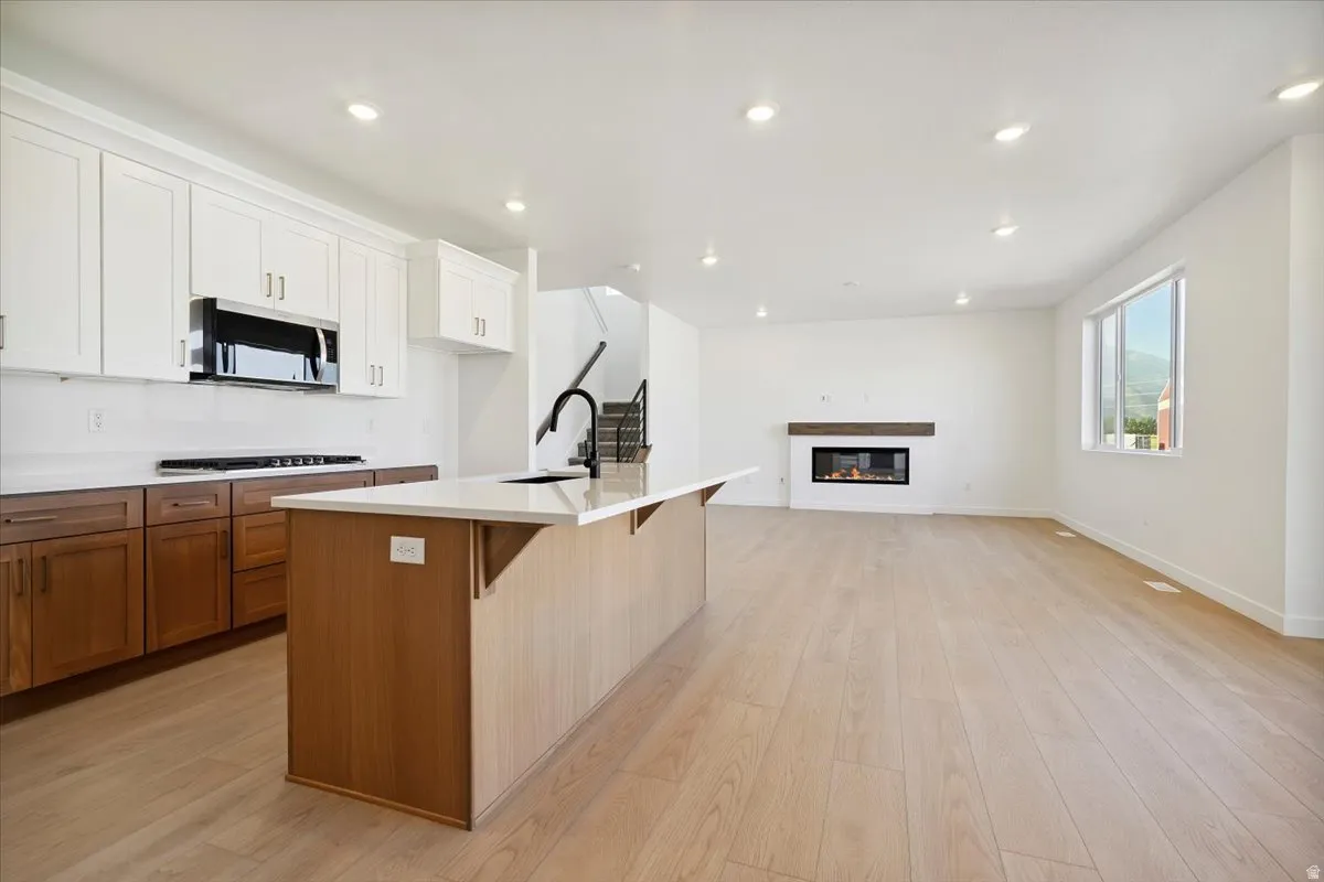 Open-concept living area featuring light wood-finish flooring, a contemporary fireplace with a wood mantel, and a kitchen island with a light-toned countertop