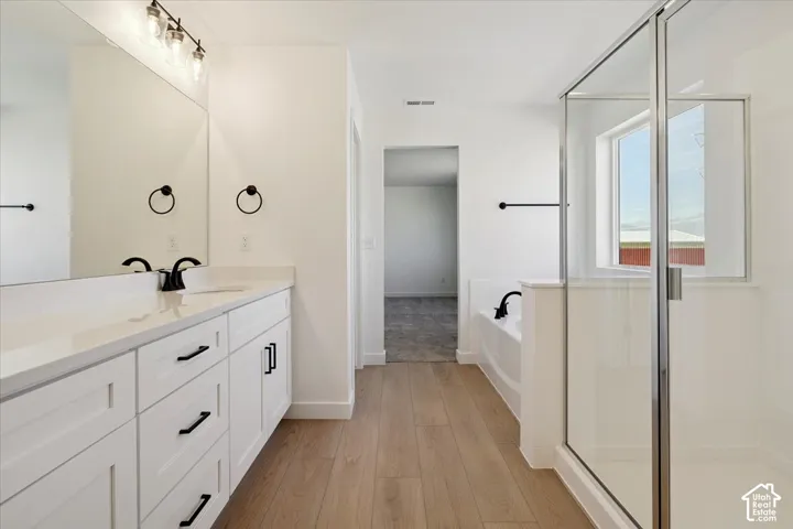 Spacious bathroom featuring a dual vanity with white cabinetry, a large mirror, a separate glass-enclosed shower, a built-in tub, and wood-finish flooring