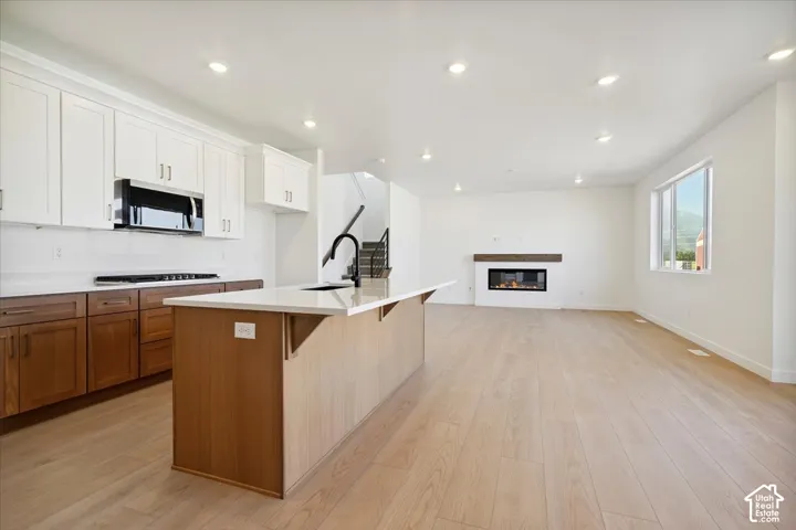 Open-concept living area featuring light wood-finish flooring, a contemporary fireplace with a wood mantel, and a kitchen island with a light-toned countertop