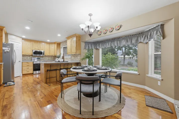Open-concept kitchen and dining area featuring wood-finish flooring, light-toned cabinetry, stainless steel appliances, a stone-finish backsplash, and a kitchen island with seating