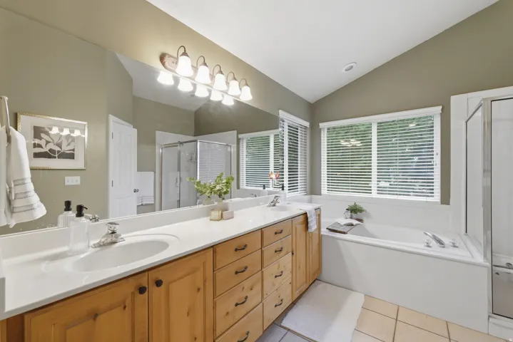 Bathroom featuring a vaulted ceiling, dual vanity with light-tone wood cabinetry, an integrated bathtub, and a glass-enclosed shower