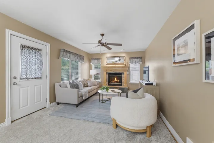 Living room featuring a wood-mantled fireplace with a stone surround, light-toned carpeting, ceiling fan, and multiple windows
