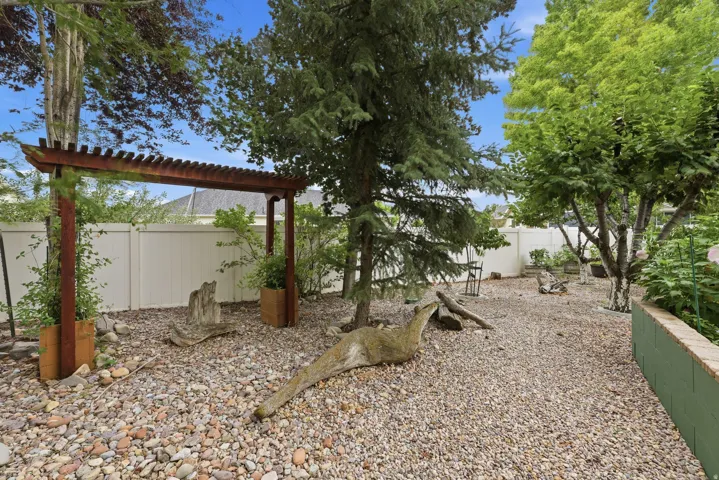 Gravel yard featuring a wooden pergola, mature trees, a privacy fence, and a brick-capped retaining wall