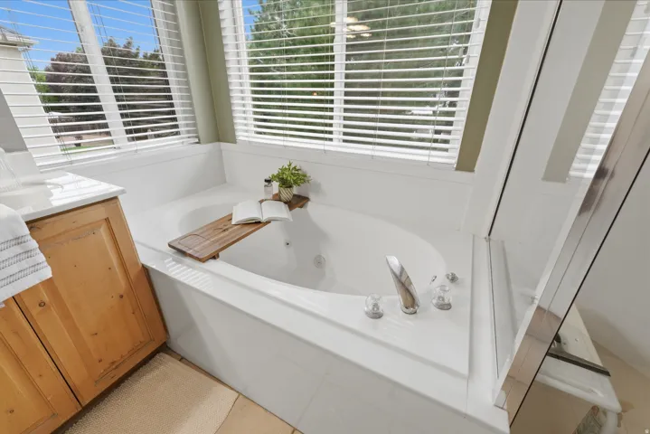 Soaking tub with jetted features, surrounded by multiple windows with blinds, chrome fixtures, and a light wood-finish vanity with a white countertop
