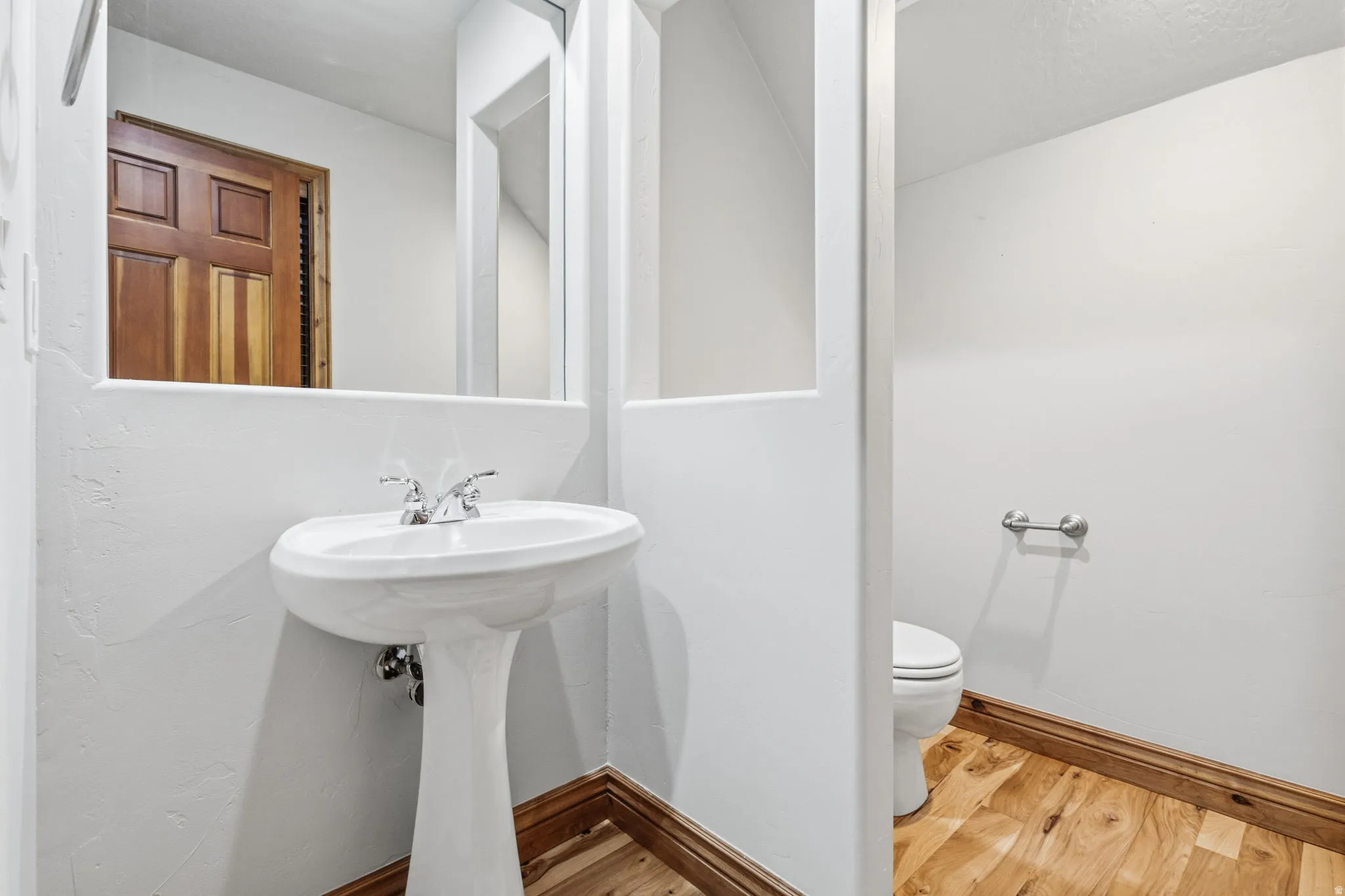 Pedestal sink with chrome faucet, wood-finish flooring, white walls, and a wood panel door