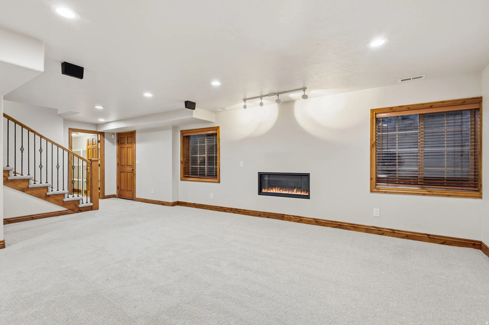 Spacious room featuring light-colored carpet flooring, an inset electric fireplace with a flame effect, and two windows with wood-frame blinds