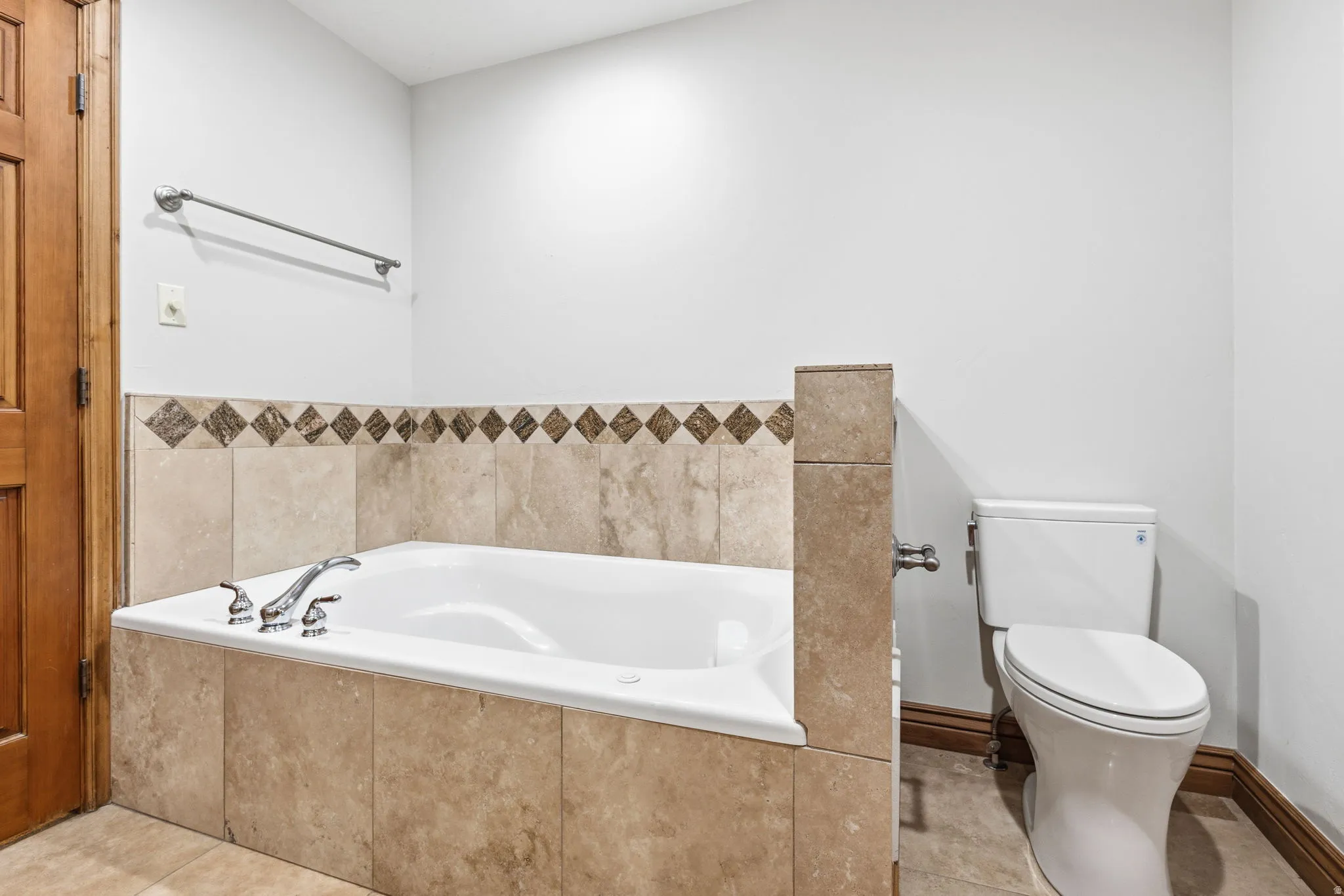 Alcove bathtub with chrome fixtures, surrounded by neutral-toned tiled skirting and a decorative diamond-patterned accent border
