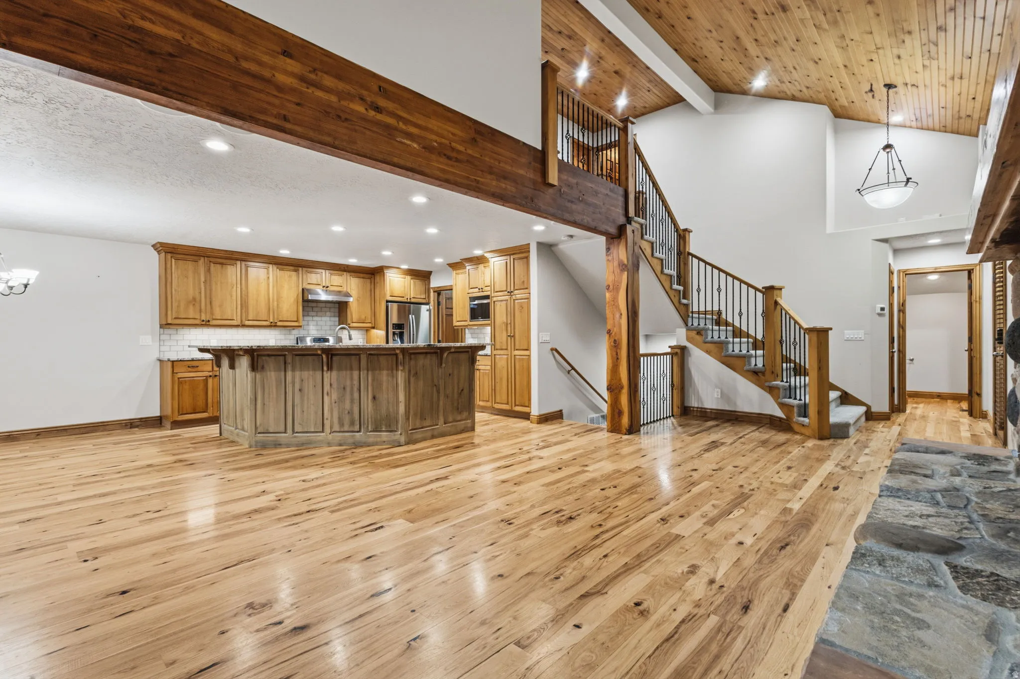Open-concept living space featuring wood-finish flooring, a kitchen with wood cabinetry and stainless steel appliances, a central island with a breakfast bar, and a staircase with wrought iron balusters