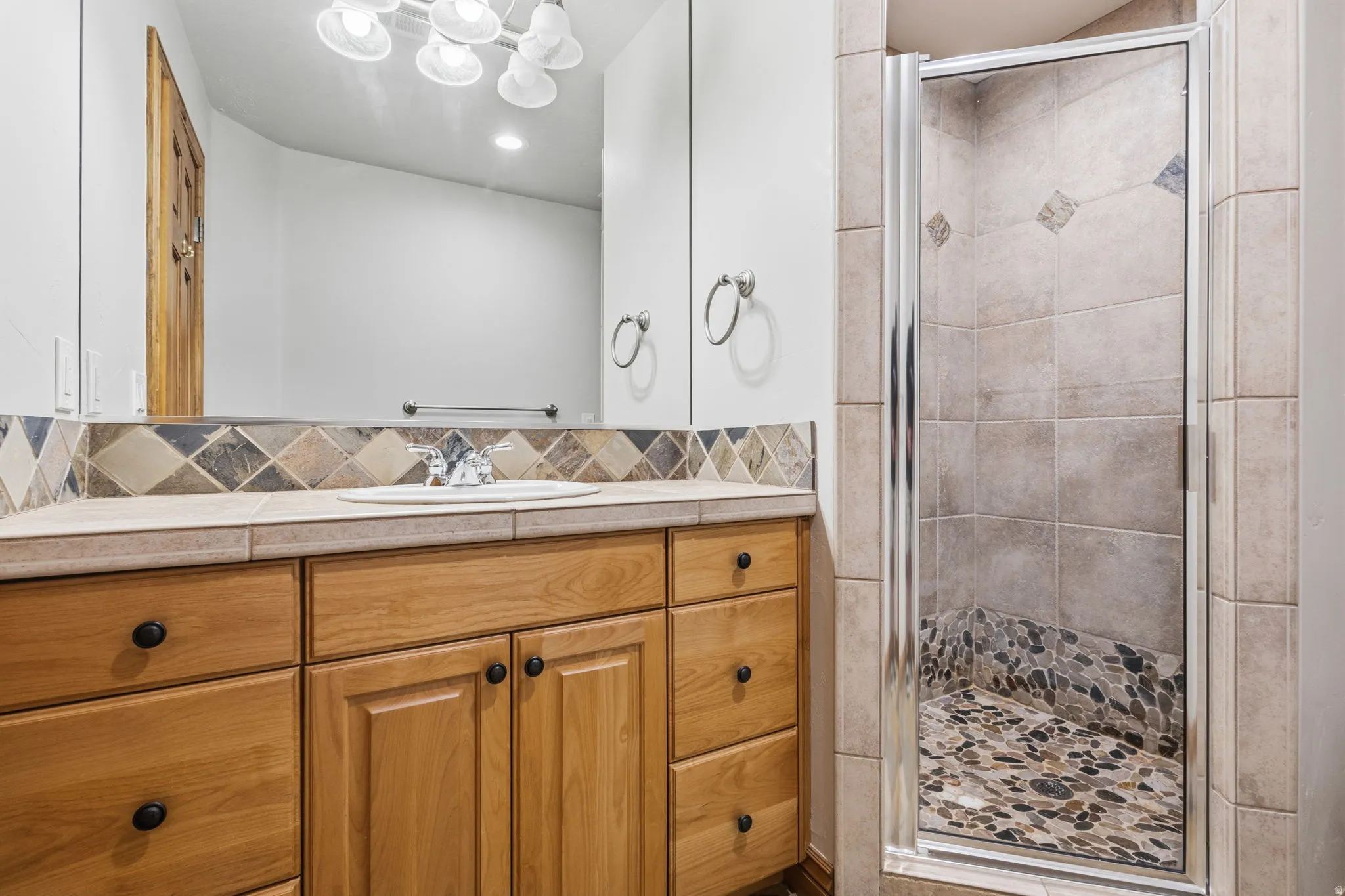 Bathroom vanity with a light wood-finish cabinet and black hardware, topped with a tiled countertop and decorative tile backsplash
