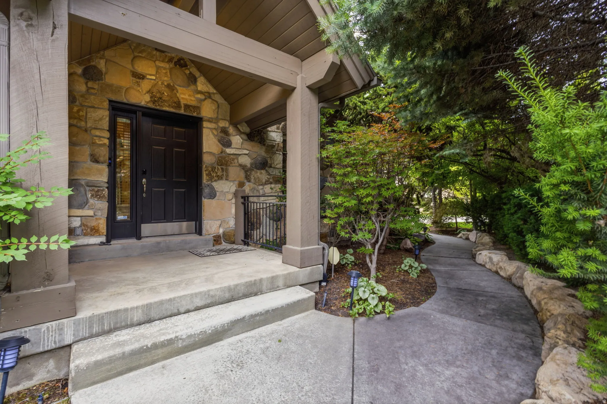 Stone facade entry with a dark paneled door and sidelight, featuring concrete steps and a covered porch