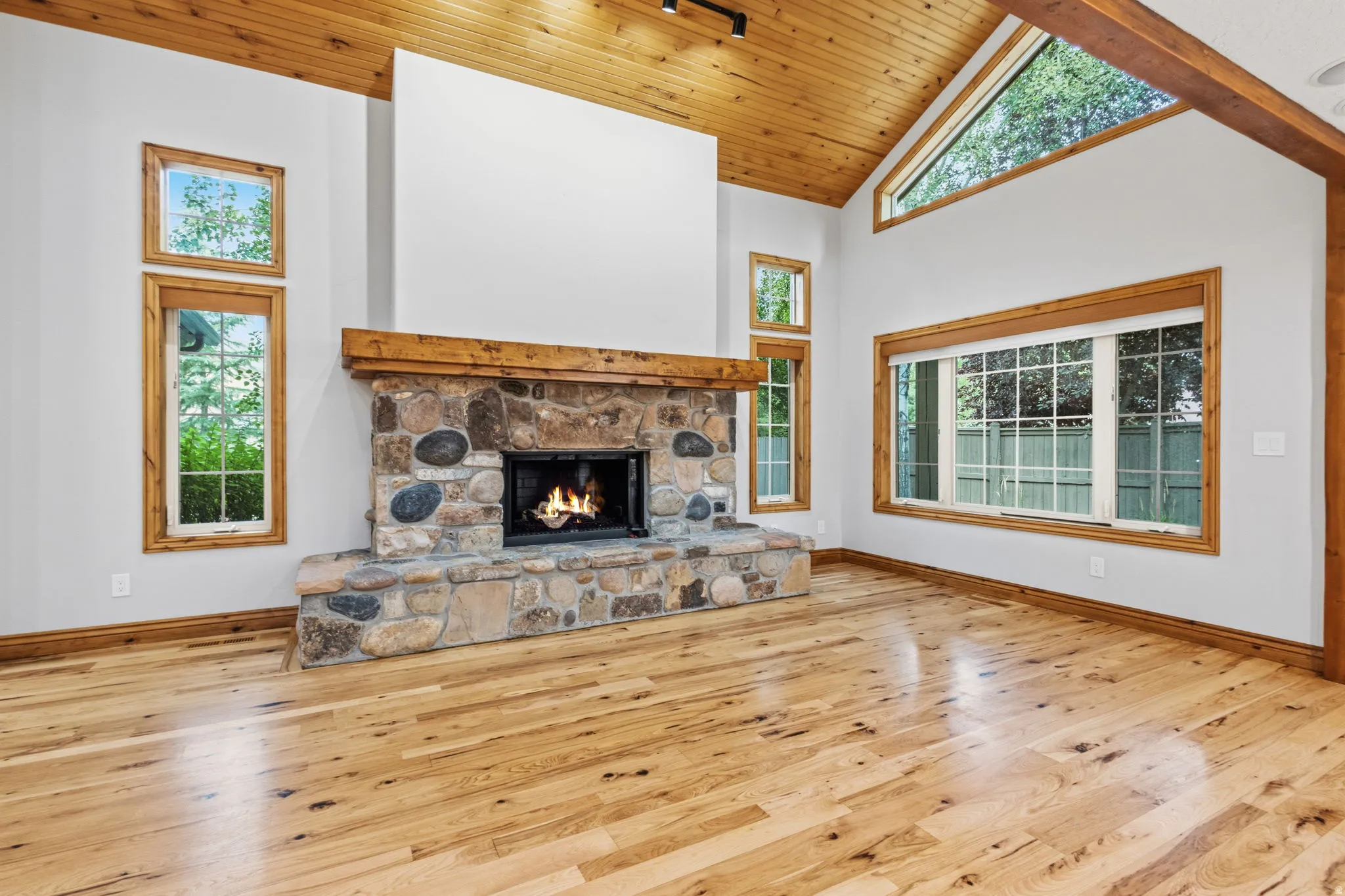 Spacious interior featuring a floor-to-ceiling stone fireplace, vaulted wood-paneled ceiling, light wood-finish flooring, and natural wood window frames
