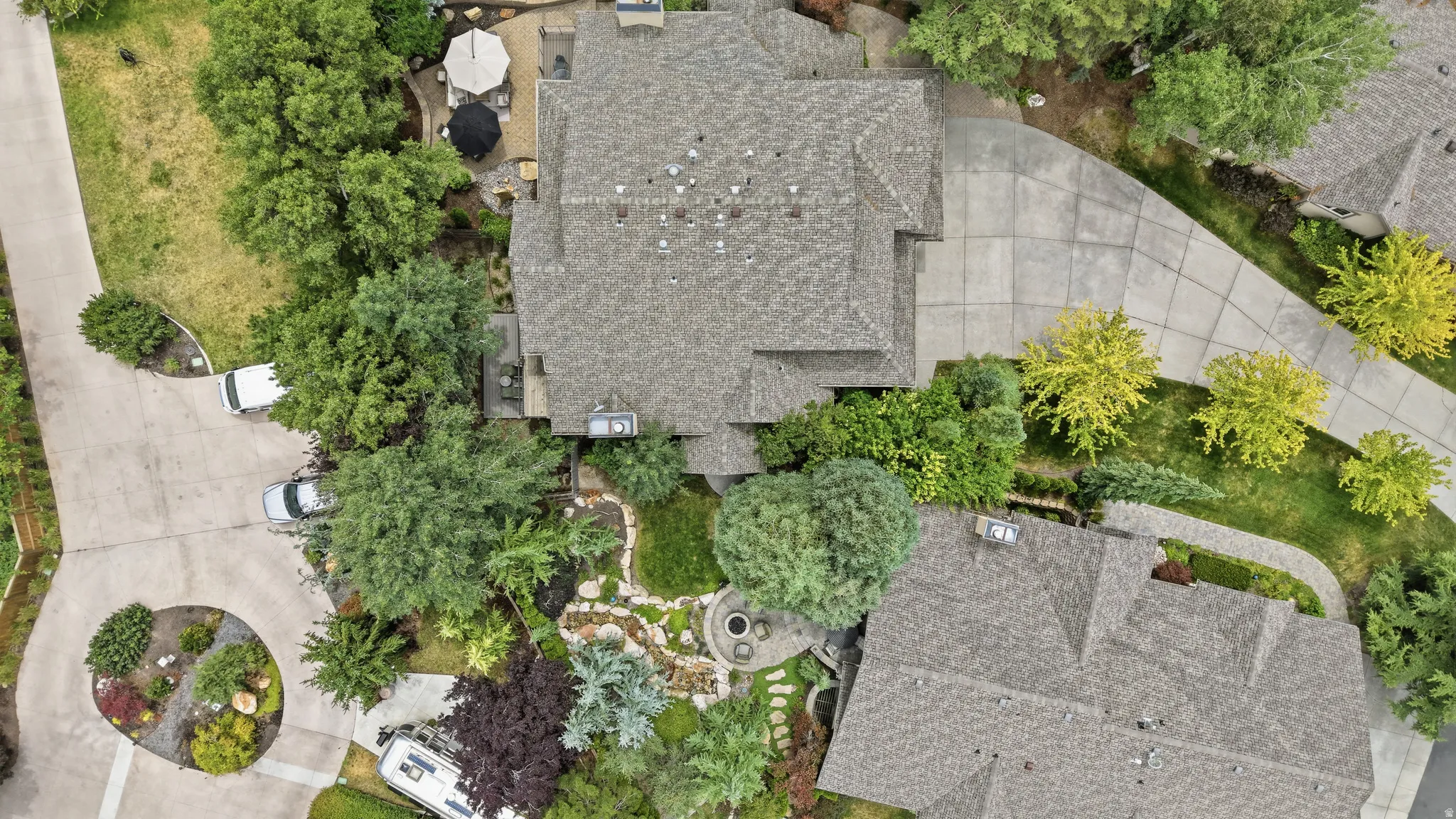 Property featuring a multi-gabled roofline, extensive paved driveways, a circular planting bed, a terraced patio, and mature landscaping