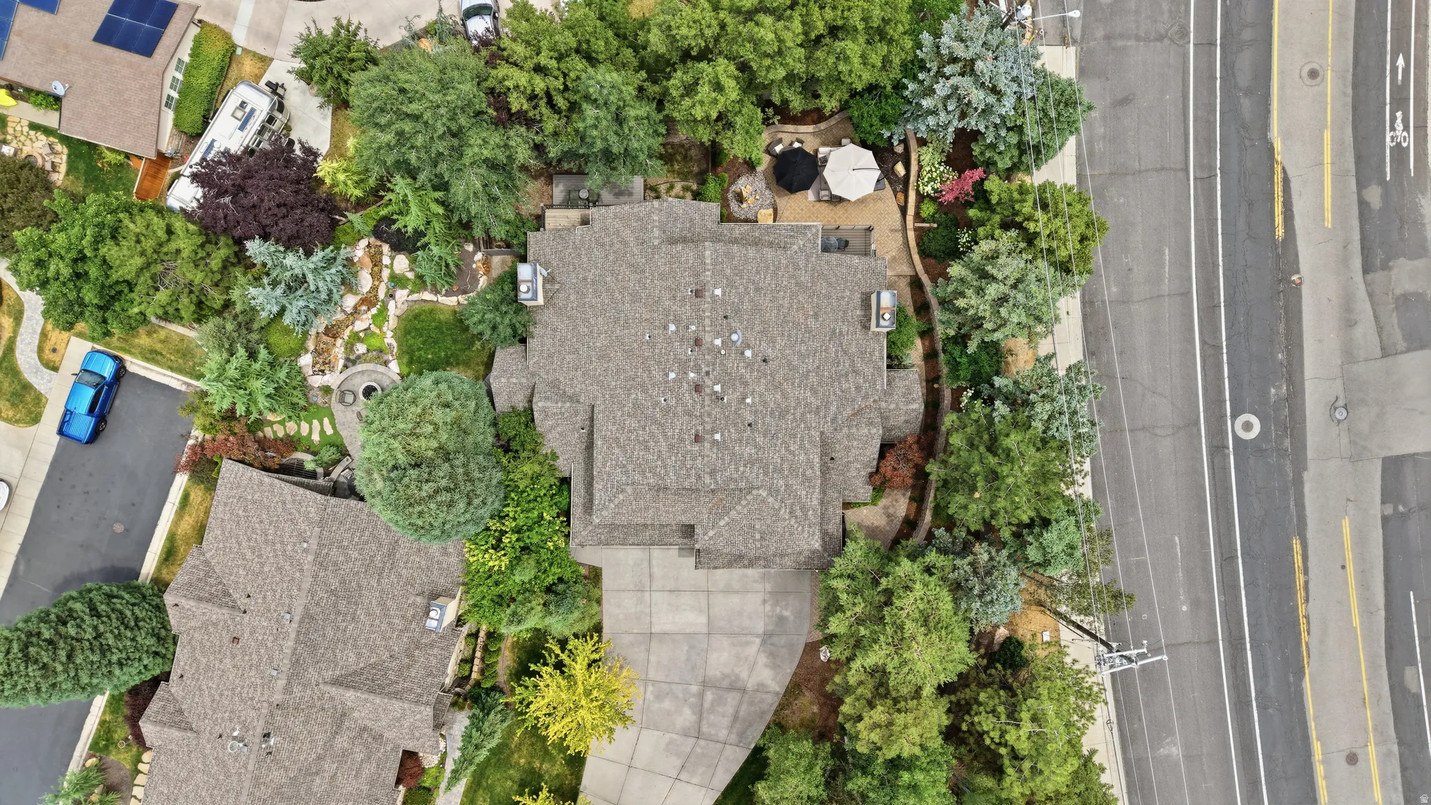 Spacious property featuring an asphalt shingle roof, paved driveway, mature trees, a landscaped patio area, and a stone walkway