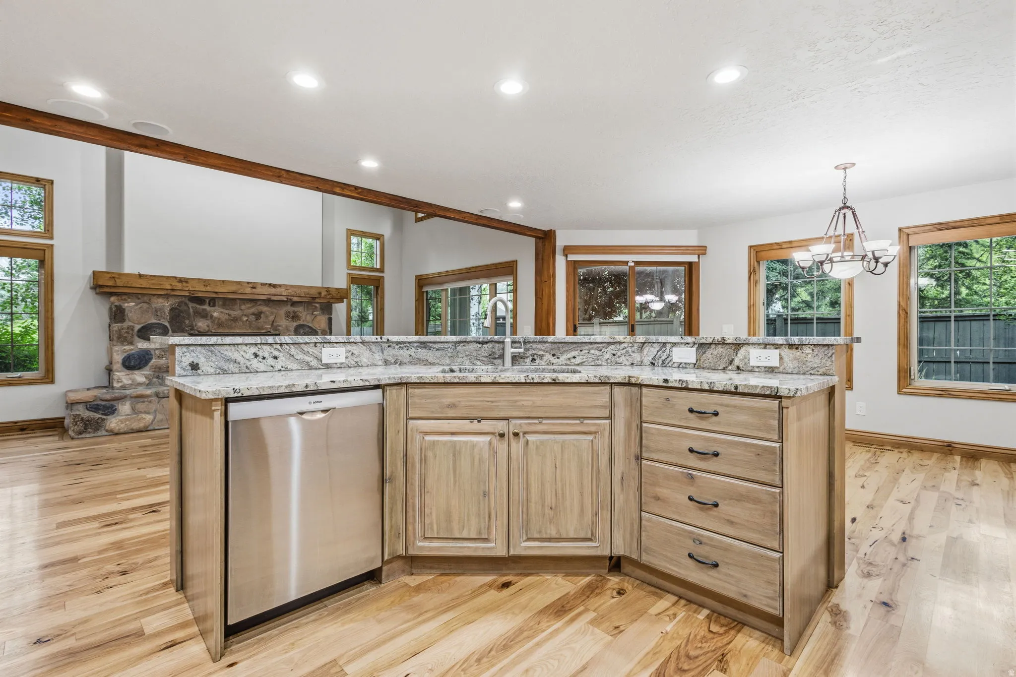 Kitchen island featuring a stainless steel dishwasher, an undermount sink, and light-toned wood cabinetry with dark hardware, all topped with a light granite countertop