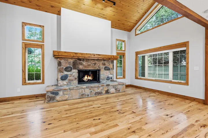Spacious interior featuring a floor-to-ceiling stone fireplace, vaulted wood-paneled ceiling, light wood-finish flooring, and natural wood window frames