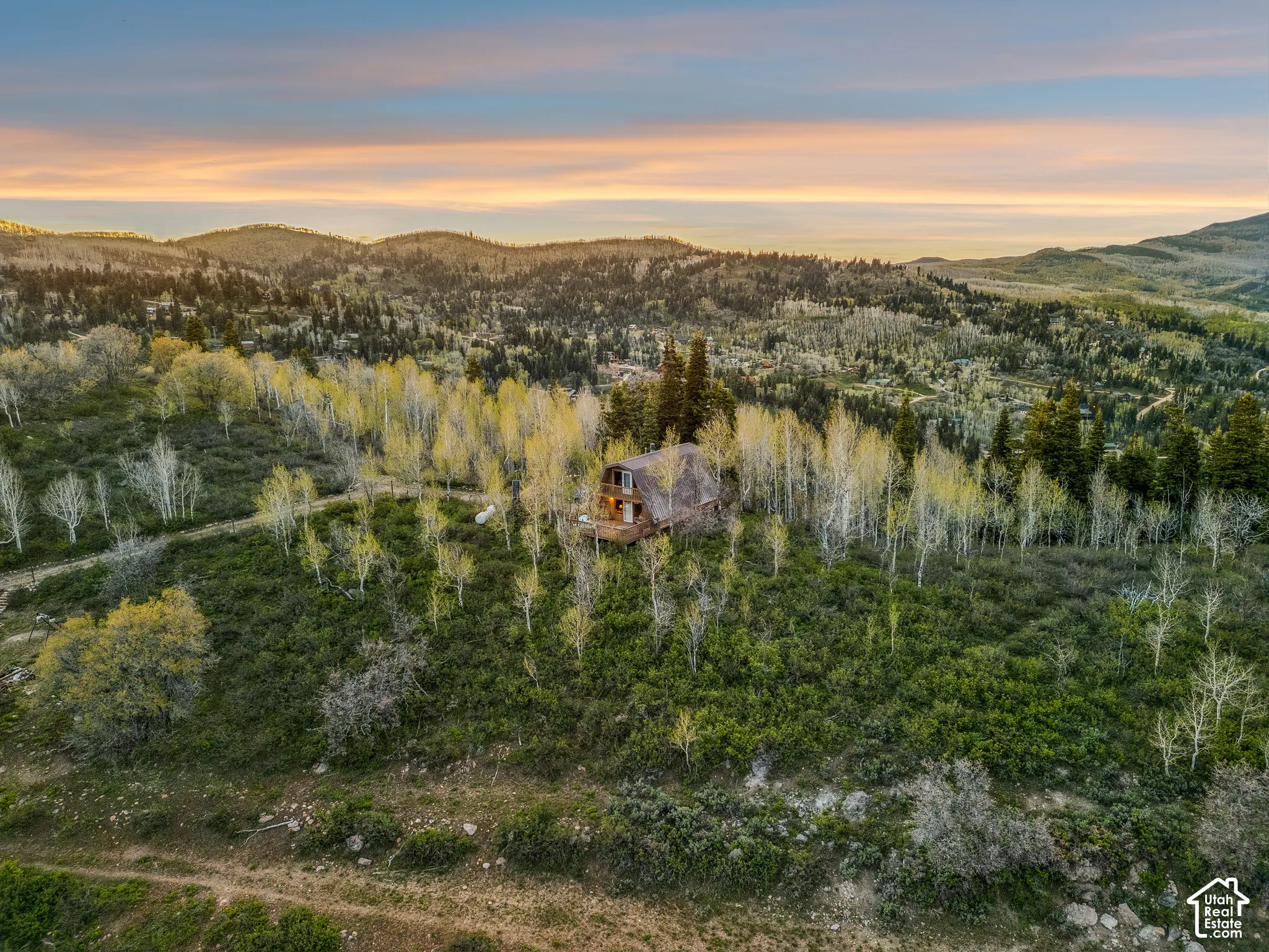 Aerial Exterior - A-frame cabin set among mature aspens and evergreens, with a standing-seam metal roof, upper balcony, and expansive wraparound deck overlooking the surrounding ridgeline.