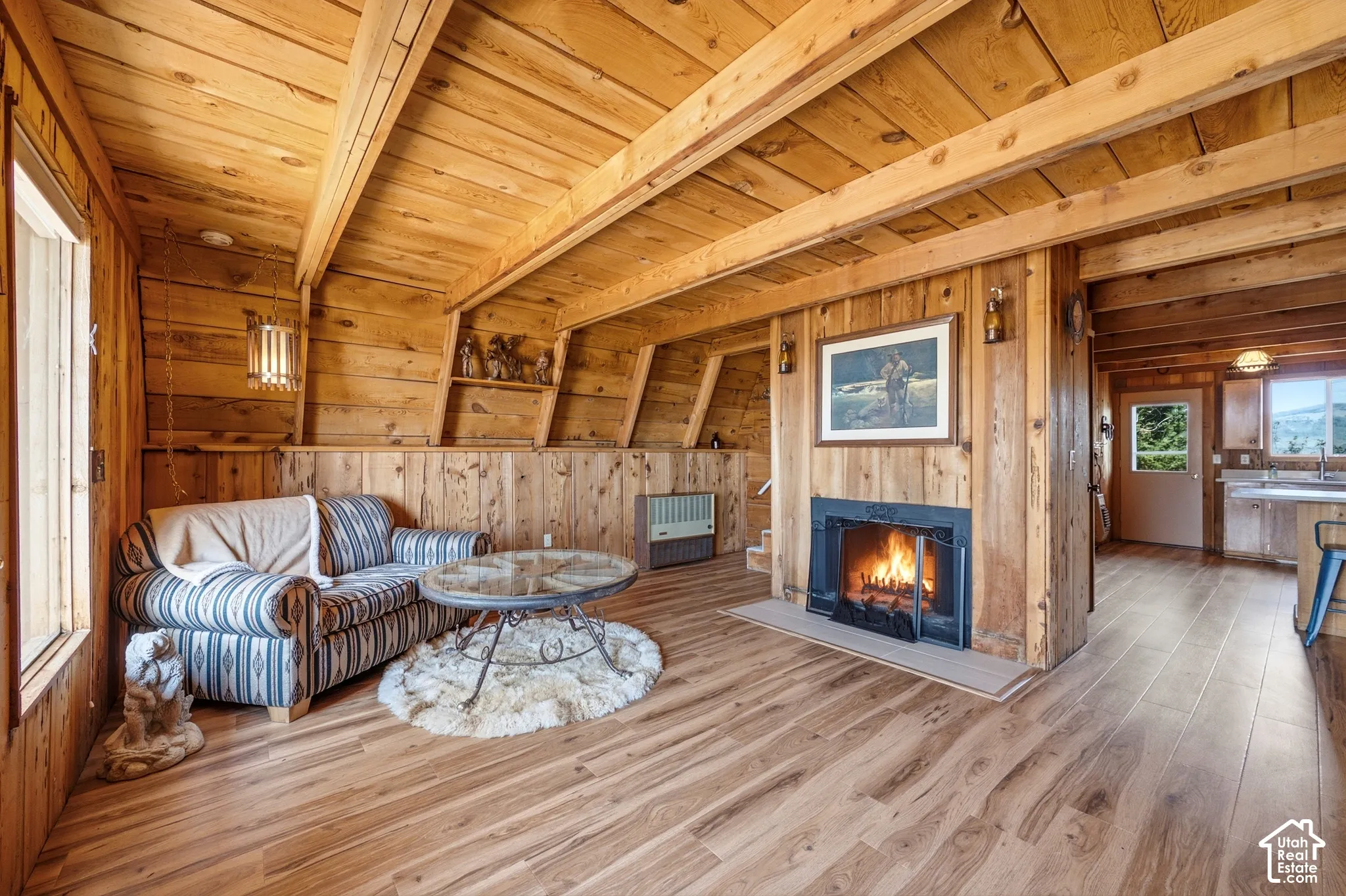 Living Area - Seating area anchored by the central fireplace, with knotty pine walls, a wall-mounted heater, and stairs leading to the upper level. Sightlines extend through to the kitchen.