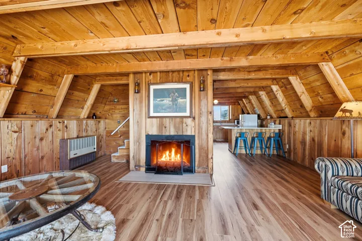 Great Room - Open living space framed by exposed wood beams and tongue-and-groove ceilings, with a wood-burning fireplace set into a full-height wood surround. Plank flooring carries through to the kitchen and dining area.