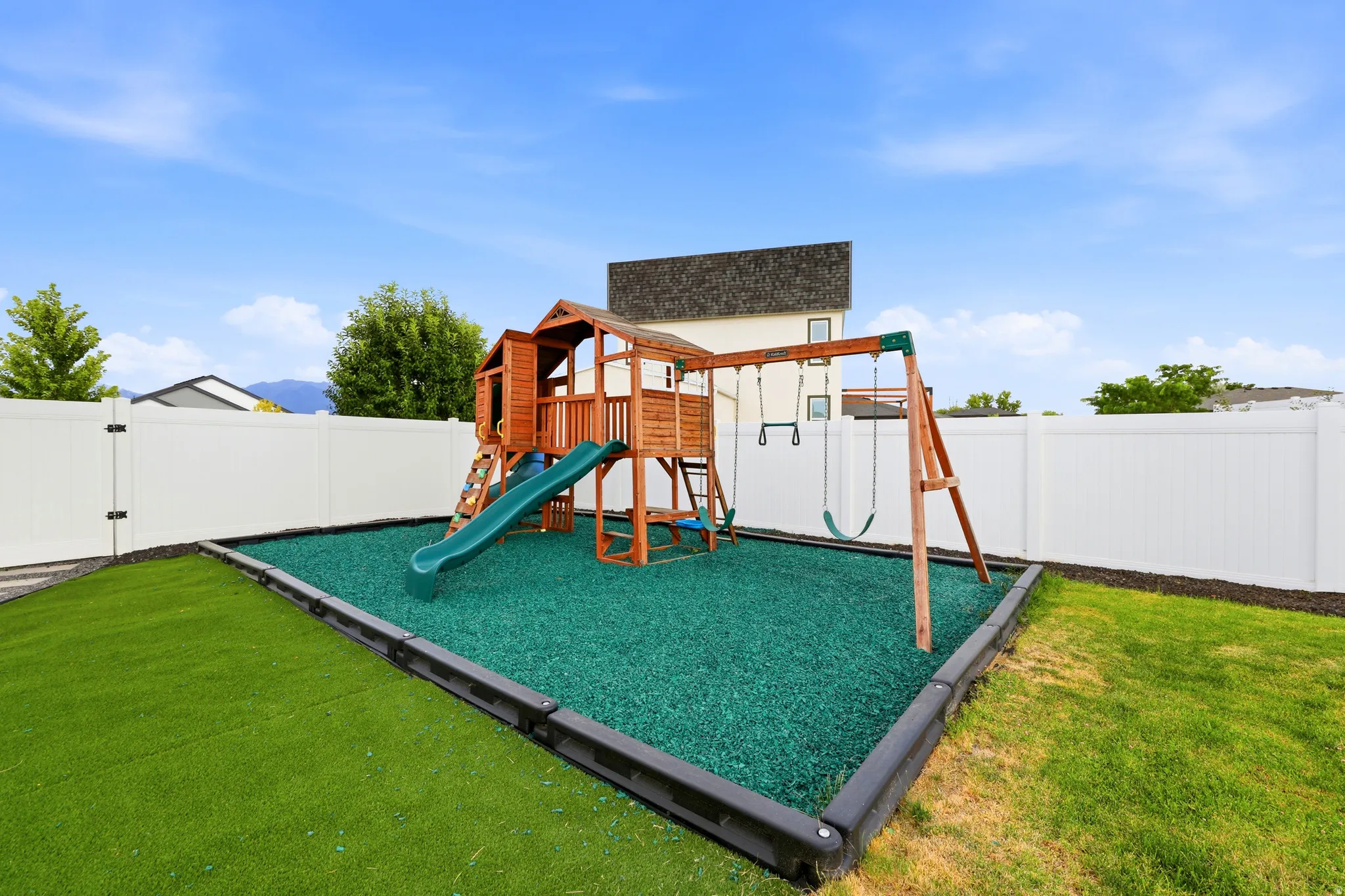 Backyard featuring a wooden playset with a slide and swing set, surrounded by green rubber mulch