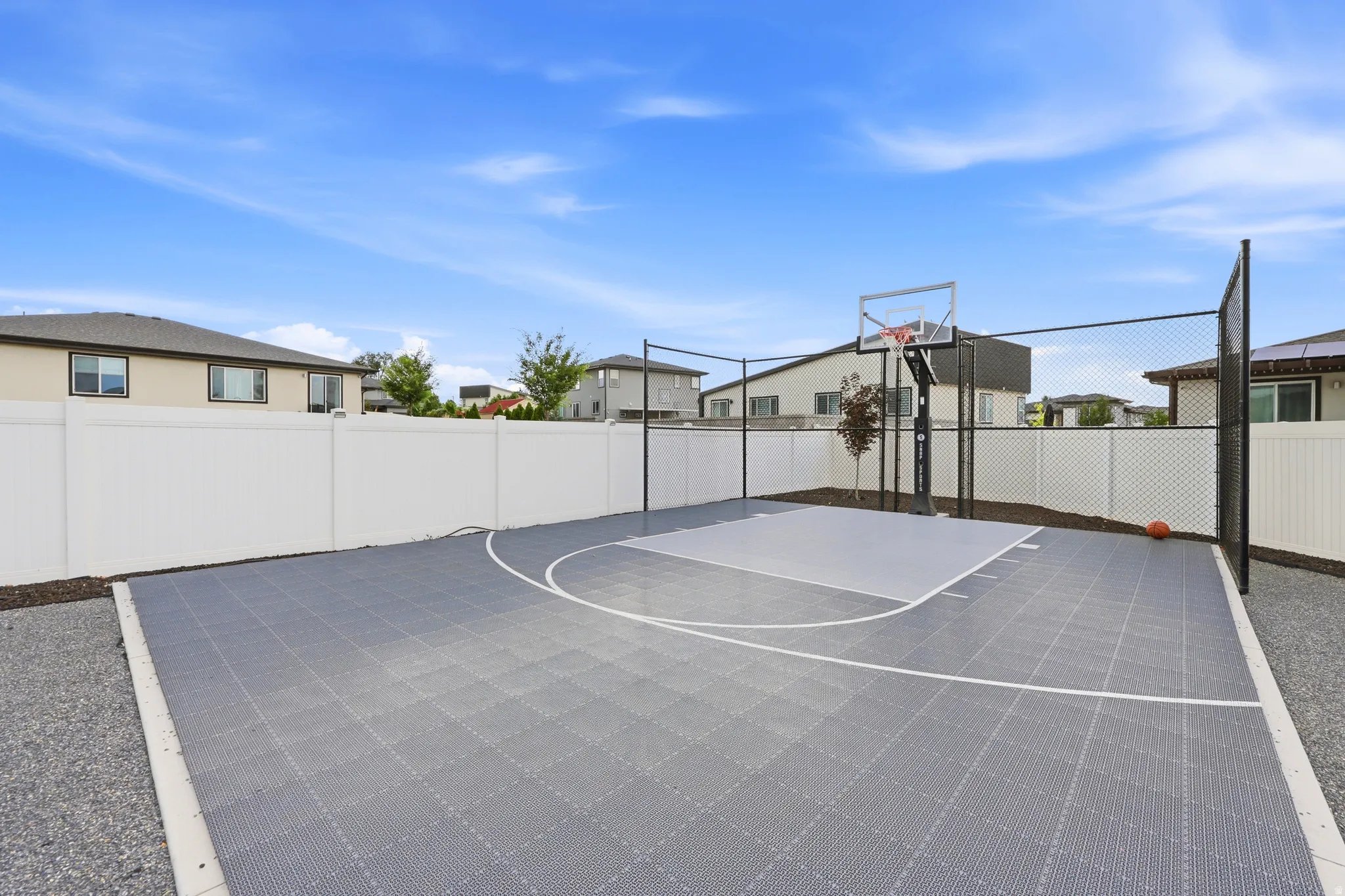 Backyard sports court featuring a half-court layout with a basketball hoop, white perimeter fencing, and surrounding gravel landscaping