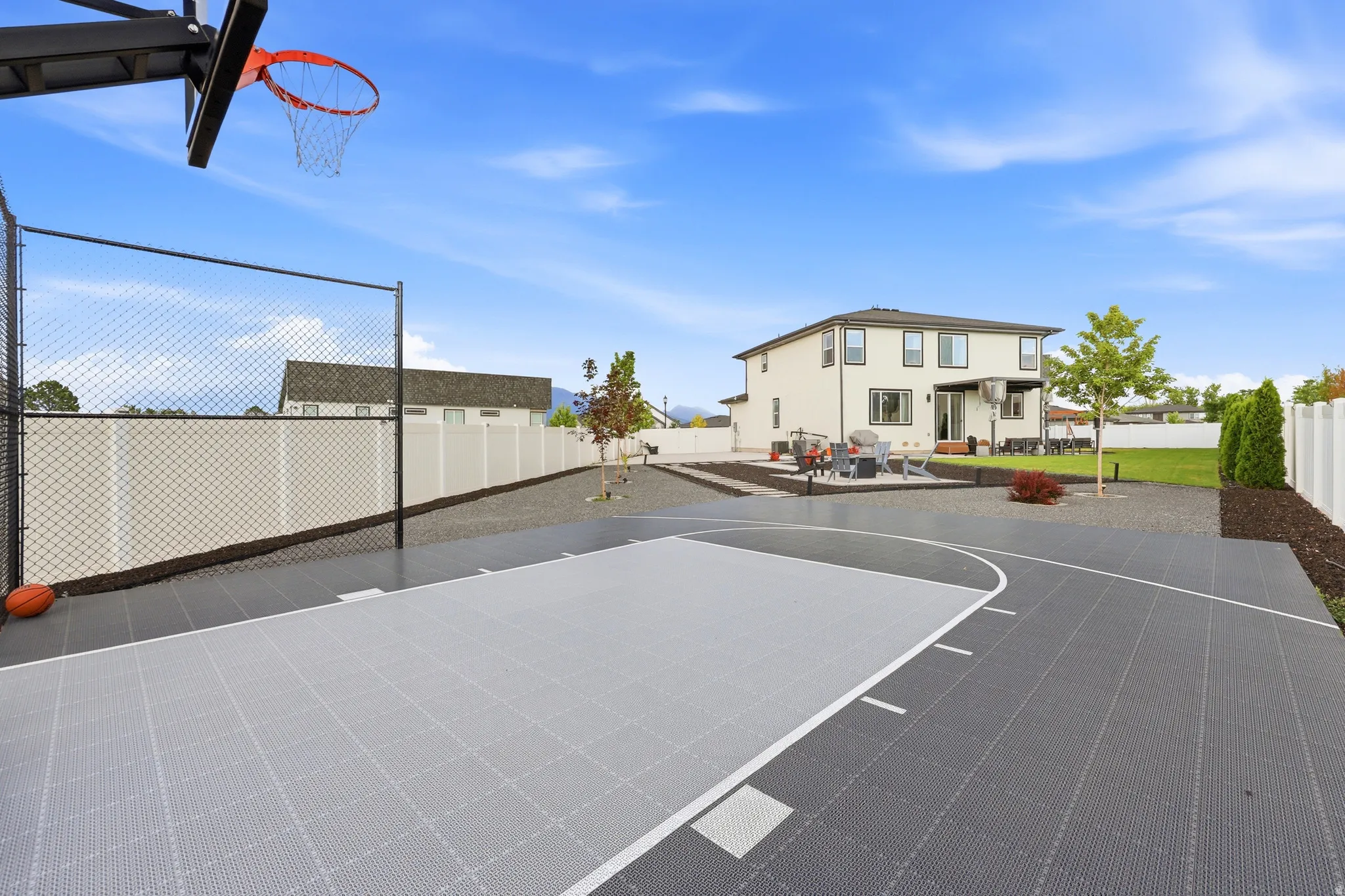 Backyard court featuring a basketball hoop with black metal support, gray and light gray playing surface, and black chain-link fencing