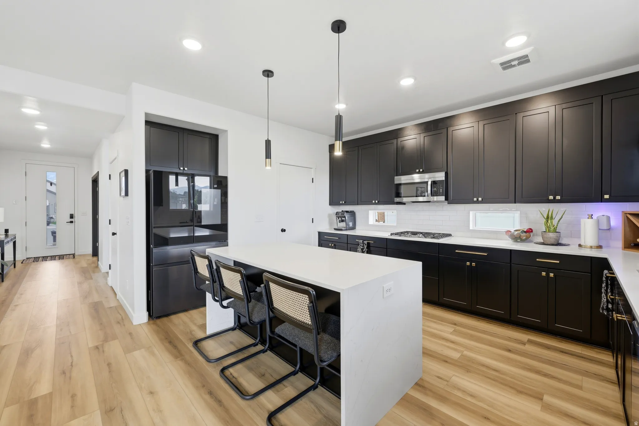 Contemporary kitchen featuring an expansive island with seating, dark shaker cabinetry, stainless steel appliances, pendant lighting, and wood-finish flooring