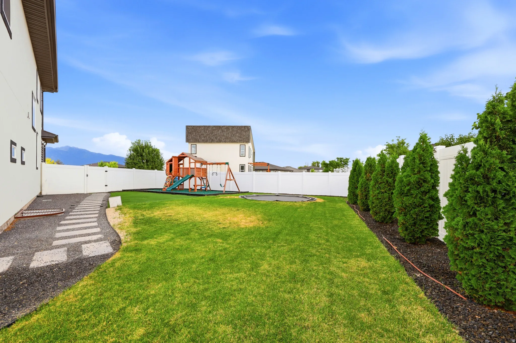 Expansive backyard featuring a large green lawn, white vinyl fencing, a stepping stone pathway, and a row of mature evergreen trees
