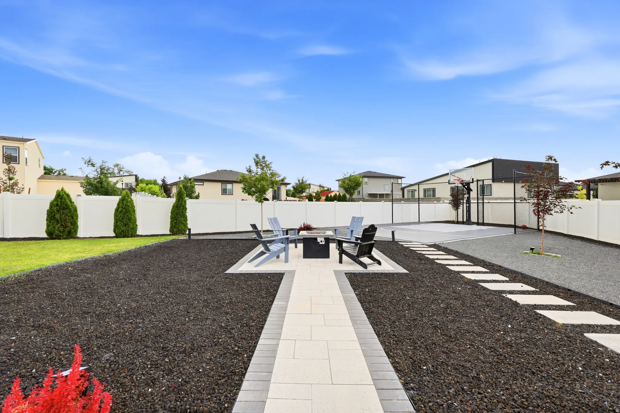Landscaped backyard featuring a paved patio area with a built-in fire pit, a dedicated sports court with a basketball hoop, and a gravel perimeter