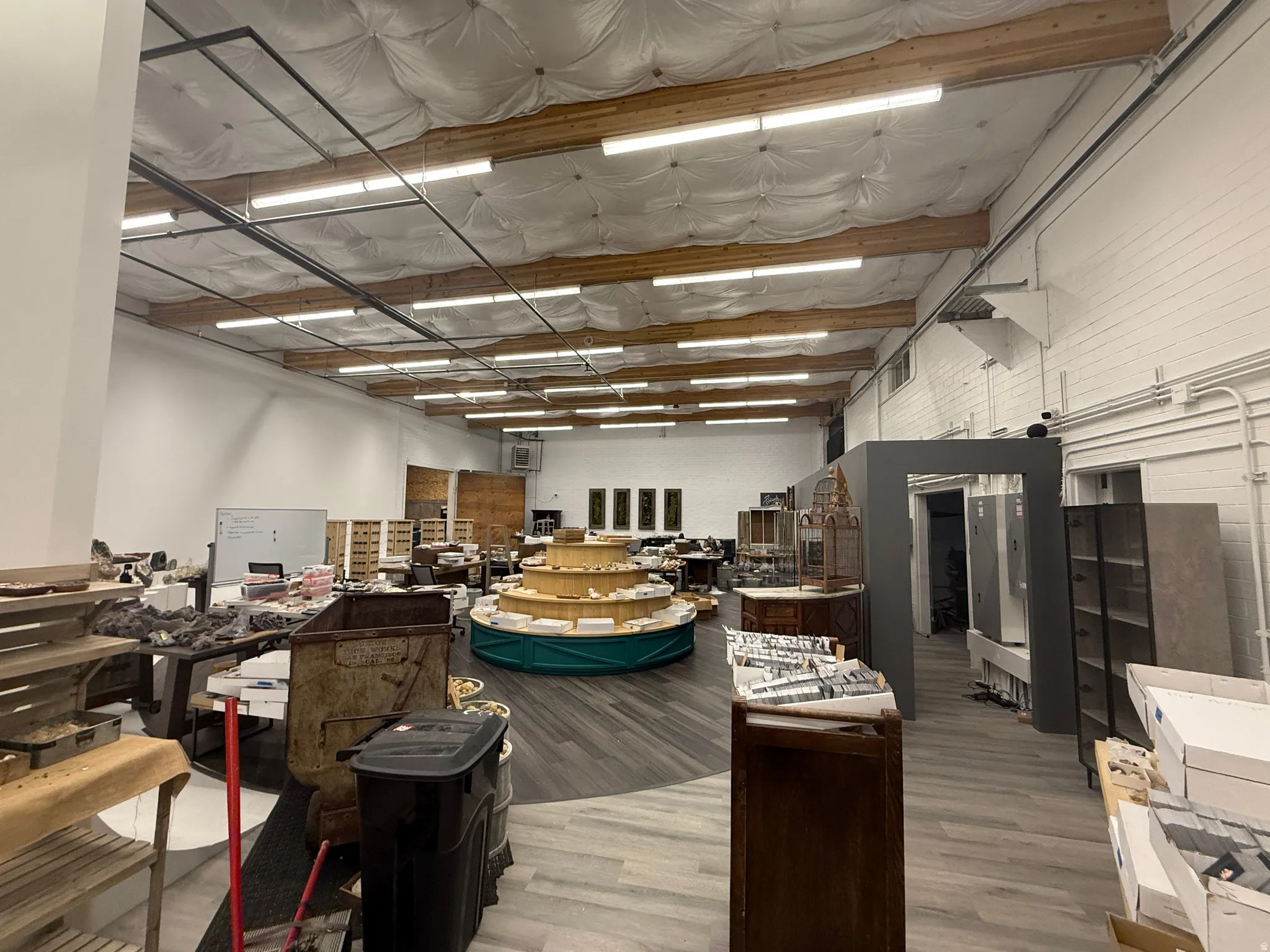 Spacious commercial interior featuring high ceilings with exposed wooden beams, extensive fluorescent lighting, white brick walls, and wood-finish flooring