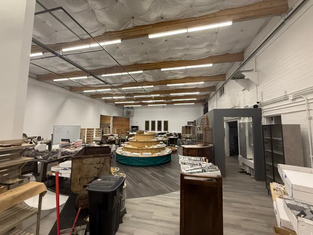 Spacious commercial interior featuring high ceilings with exposed wooden beams, extensive fluorescent lighting, white brick walls, and wood-finish flooring